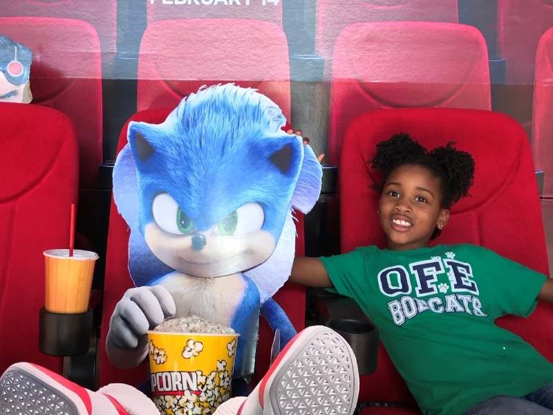 Picture of a cardboard Sonic the Hedge Hog and Harper L Ferguson at the movie theater sitting in a chair.